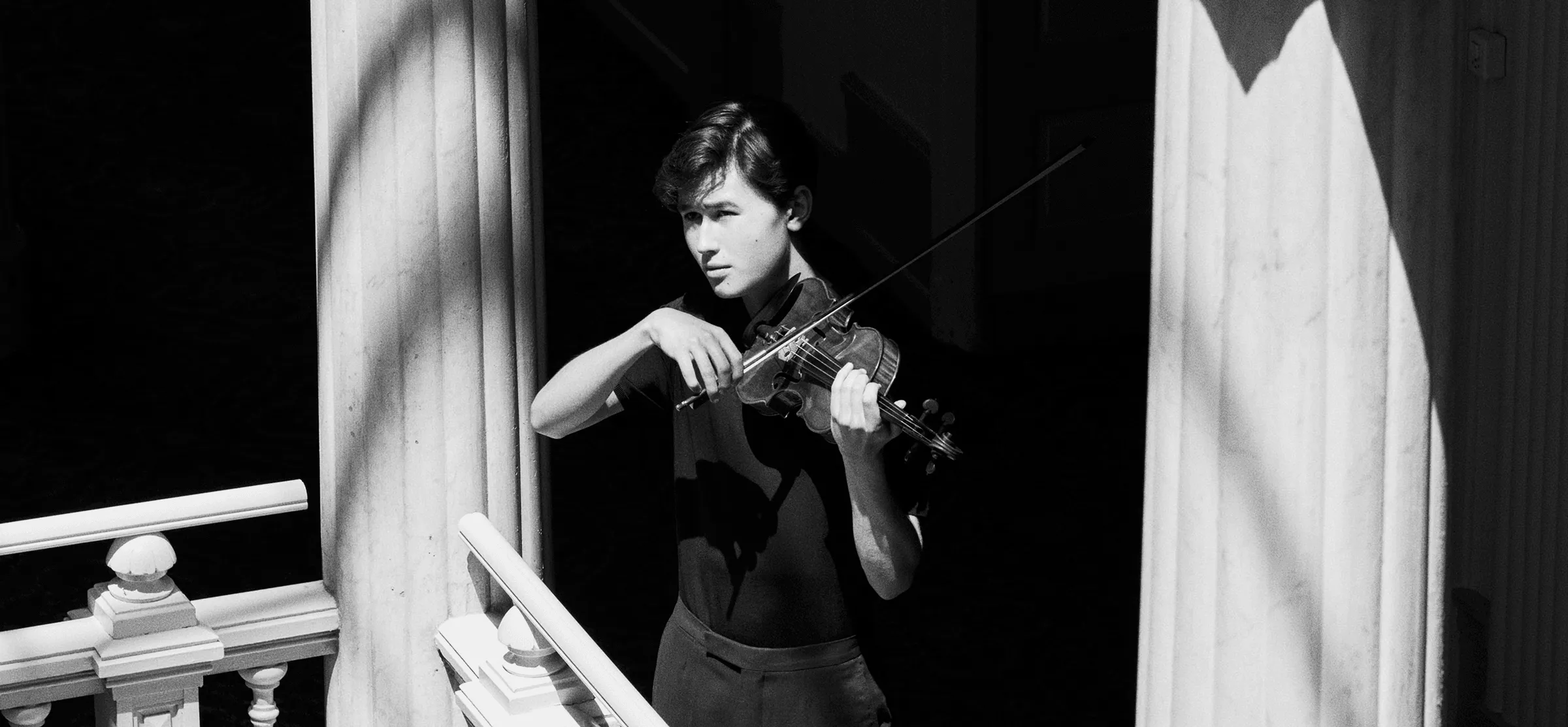 Foto em preto e branco do jovem violinista Daniel Lozakovich. Ele está segurando um violino entre duas colunas com o sol batendo em seu rosto.