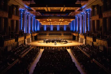 Sala de concertos com pessoas na plateia e no palco. As colunas estão iluminadas de azul e o palco com uma luz quente.