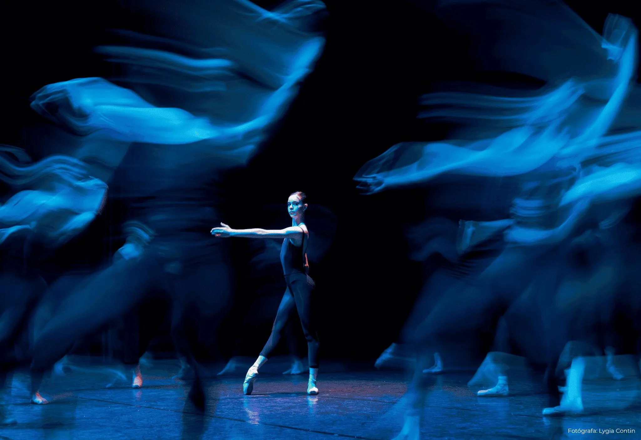 Uma dançarina parada em posição no centro da imagem, demais dançarinos da Cia. Ballet Paraisópolis em volta em movimento