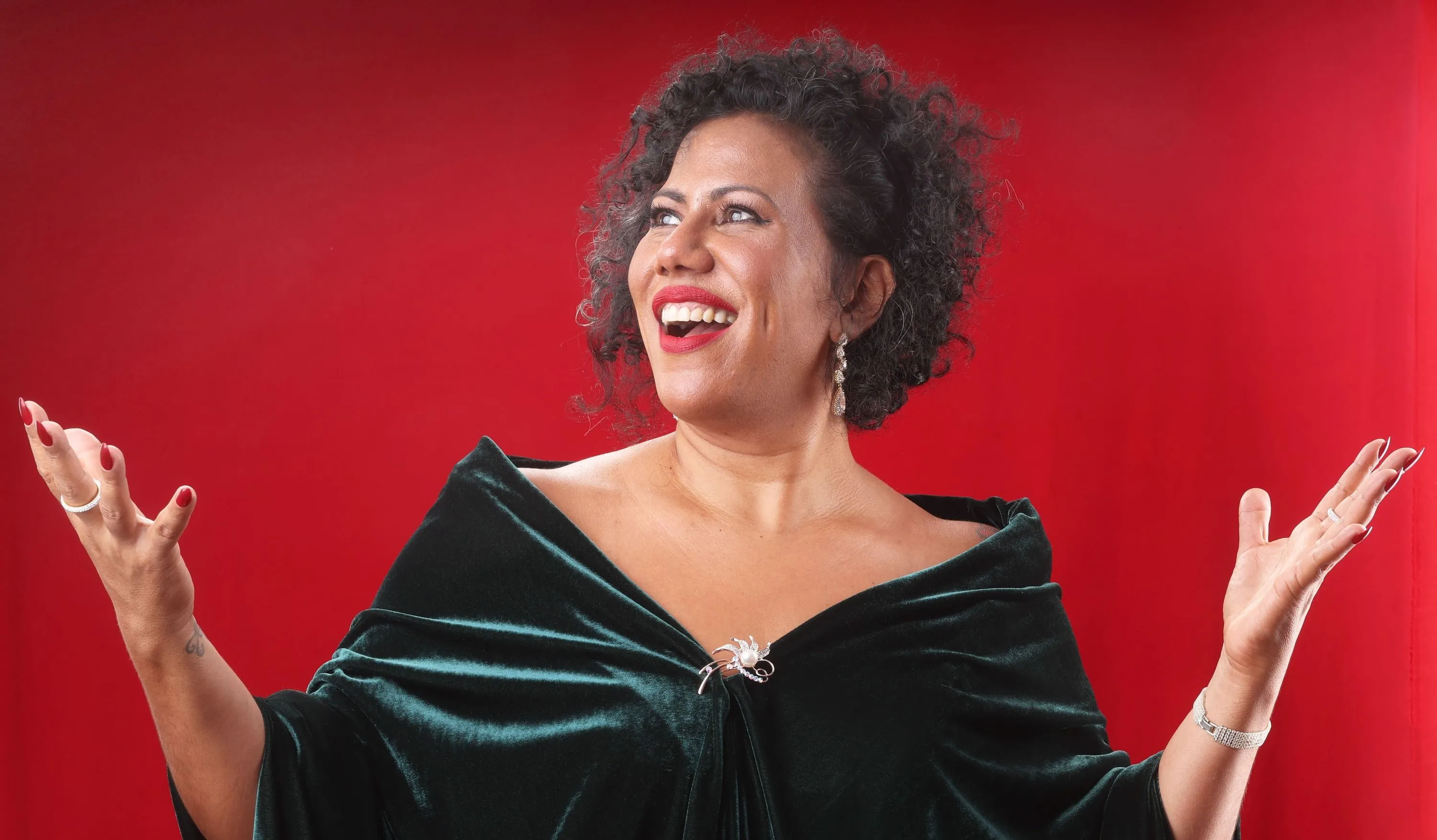 Indiana Nomma é uma mulher negra de pele clara com cabelos curtos cacheados. Está olhando para o lado em um fundo vermelho.