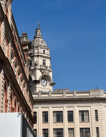 Foto do lado externo da Sala São Paulo, no sentido do estacionamento. Em destaque, a torre do relógio, onde está hasteada a bandeira da cidade. Do lado direito, parte do prédio laranja da Pinacoteca e ao fundo o céu azul num dia ensolarado.