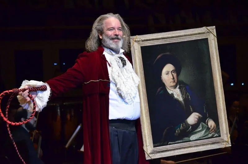 Foto do ator Jairo Mattos, homem branco, de barba e cabelos longos e grisalhos. Eles veste uma roupa de gala antiga, com camisa branca bufante e um casaco vermelho. Segura um quadro em sua mão.