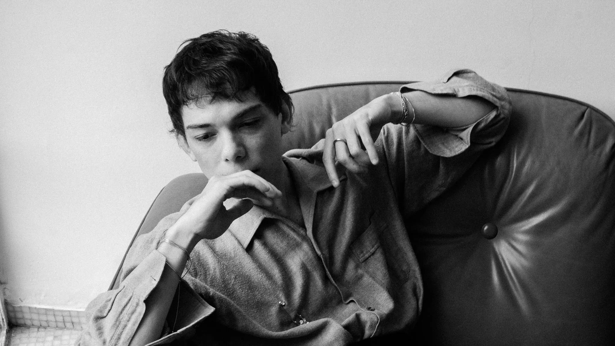 Foto em preto e branco de Vitor, rapaz branco de cabelo curto e liso, sentado em um sofá de couro