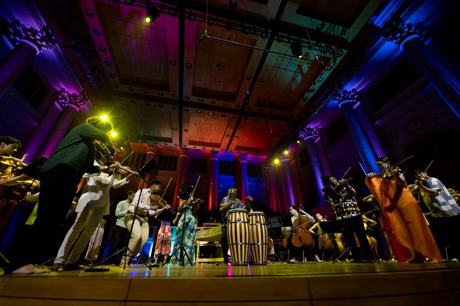 Grupo de músicos, com percussão e instrumentos de cordas, tocam de pé no palco da sala sao paulo. O espaço é iluminado por luzes coloridas.