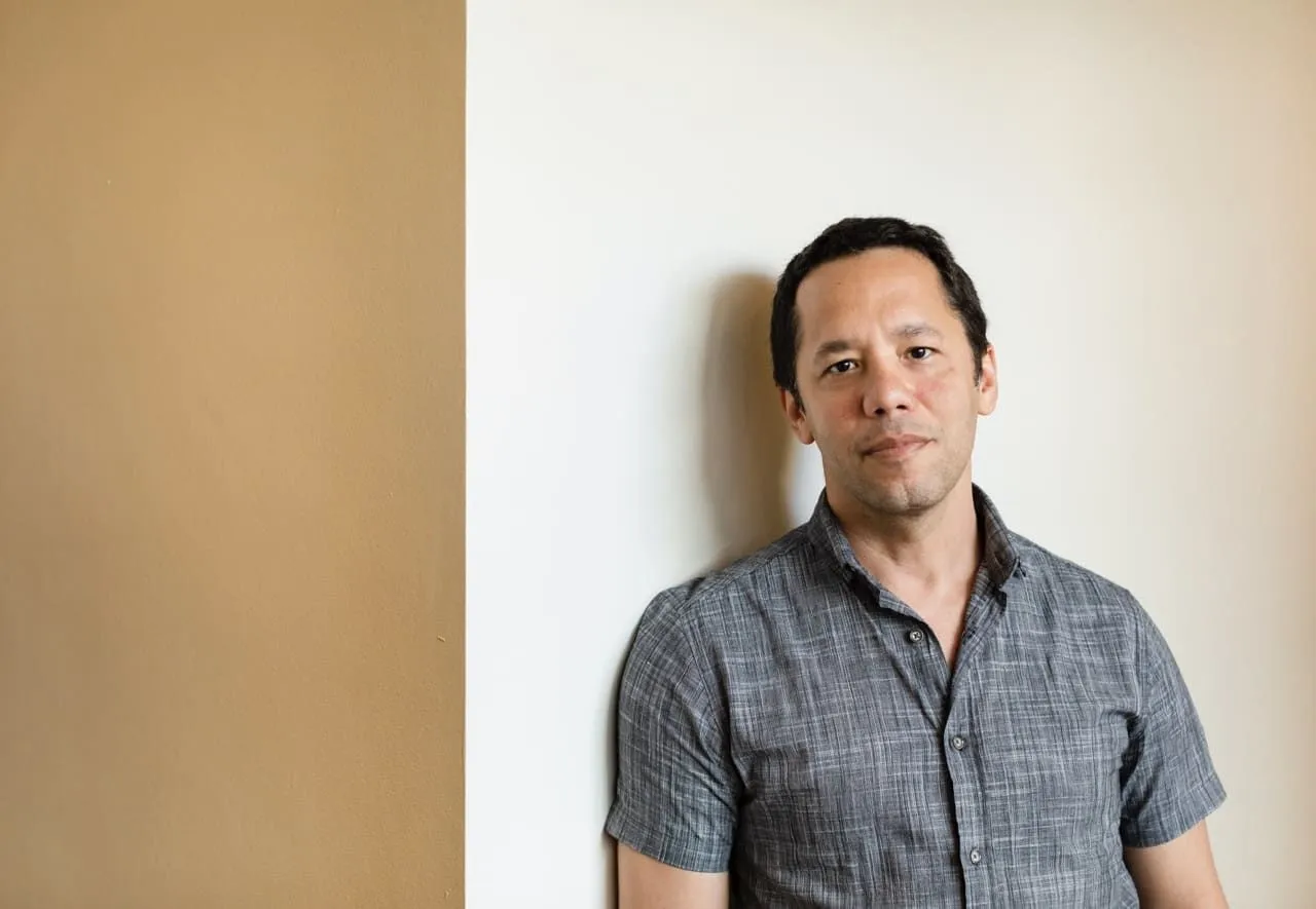 Autor Itamar Vieira Junior com camisa xadrez cinza em parede branca e bege.