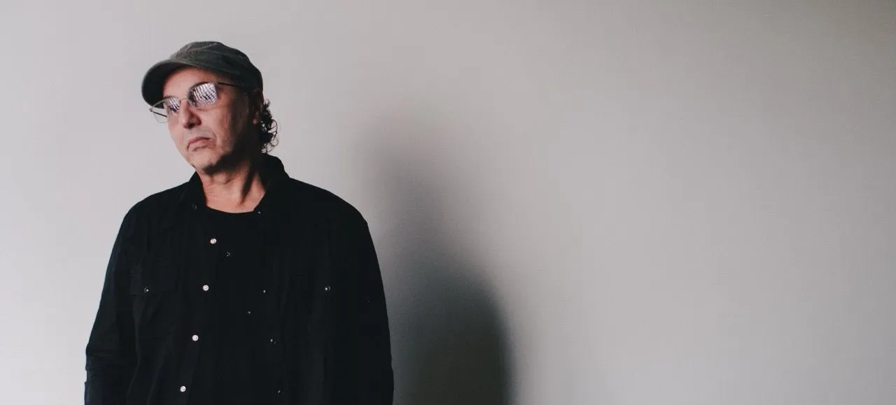 Zeca baleiro é um homem branco, usa óculos e está com uma feição séria em um fundo branco.