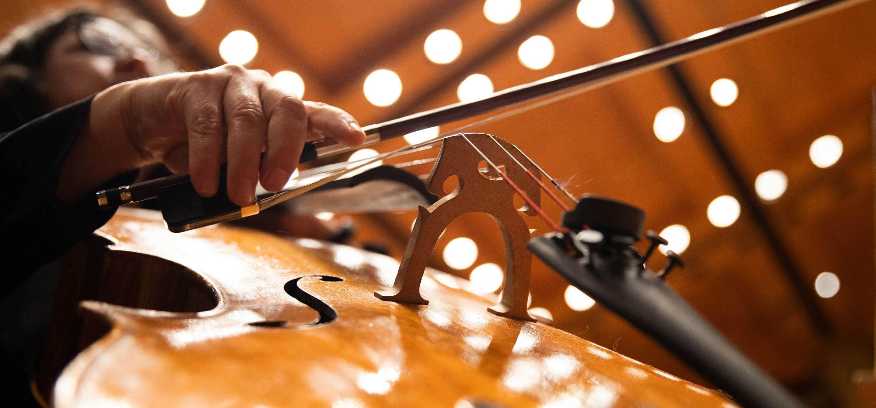 A imagem mostra um violoncelo em um ângulo de baixo pra cima, a mão do músico segurando o arco e as luzes do teto da Sala São Paulo.