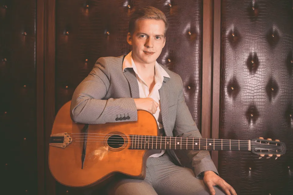 Olli Soikkeli é um homem branco de cabelos curtos e loiros, está segurando um violão e vestindo camisa clara com terno cinza.