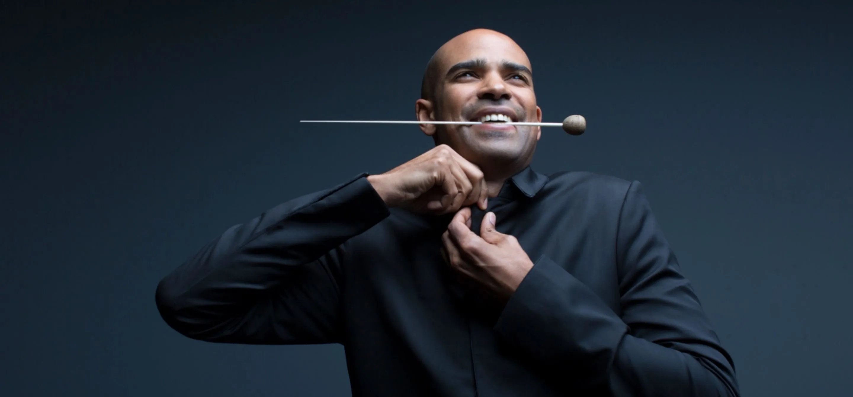Retrato de Kevin John Edusei, um homem negro e careca, mordendo sua batuta